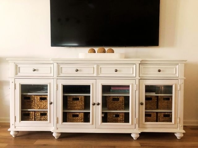 A white entertainment cabinet with glass doors neatly housing wicker storage bins, offering an elegant solution for hiding clutter under a mounted TV.