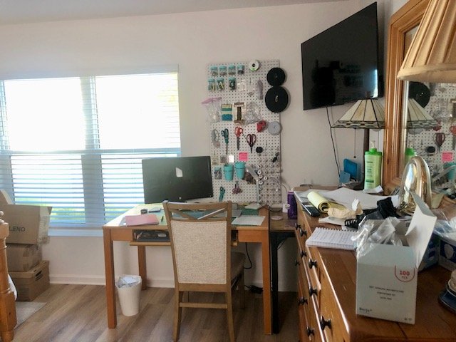 Cluttered desk in a guest room with scattered papers, craft supplies, and boxes, showing an unorganized home office space.