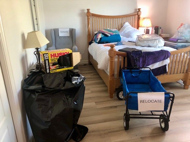 Decluttering staging area with a labeled “Relocate” wagon and large trash bags for discarded items, set beside the guest bed.